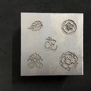 Puede incluir: Un sello de metal con cinco diseños diferentes: una hoja, una flor, el símbolo Om, un patrón paisley y un mandala.