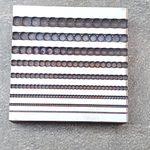 Puede incluir: Una herramienta de metal con filas de pequeños agujeros redondos. Los agujeros están dispuestos en una cuadrícula y son de diferentes tamaños. La herramienta probablemente se utiliza para crear joyas u otros objetos pequeños.