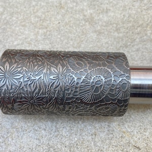 Könnte beinhalten: Ein Nudelholz aus Metall mit einem Blumenmuster. Das Nudelholz hat an jedem Ende einen glatten Griff.