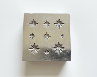 May include: A square, silver metal stamp with nine star-shaped impressions of varying sizes. The stars are evenly spaced across the surface, creating a symmetrical pattern. The stamp is likely used for jewelry making or other crafts.