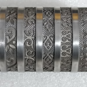 May include: A silver-colored metal tool with a series of decorative bands. Each band features a different engraved floral or ornamental pattern. The tool is mounted on a silver rod, and the bands are separated by silver rings.