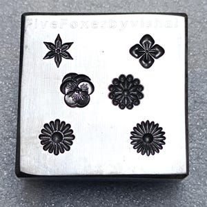 Könnte beinhalten: Ein quadratischer Metallstempel mit sechs verschiedenen floralen Designs. Die Designs umfassen einen Stern, eine vierblättrige Blume, ein Stiefmütterchen und drei gänseblümchenartige Blumen. Der Stempel trägt oben den Text "FiveFoxerbyVishel".