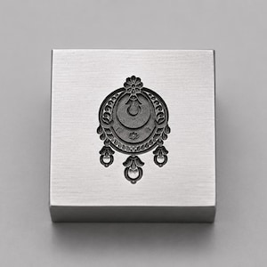 May include: A square silver-toned metal block featuring a centered, recessed black engraving of a circular pendant design. The design includes a top flower motif, a central crescent shape, and three small dangling ornaments at the bottom edge.