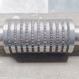 Könnte beinhalten: Eine silberne Metallrolle mit einer Reihe von sich wiederholenden Mustern, die in ihre Oberfläche graviert sind. Die Rolle hat zwei glatte Metallenden.