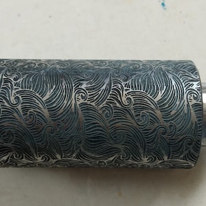 Op de afbeelding: Een zilveren metalen roller met een decoratief patroon van golven en swirls. De roller wordt gebruikt om patronen in klei of andere materialen te creëren.
