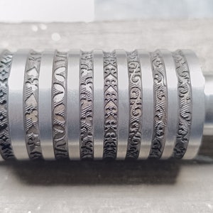 Könnte beinhalten: Eine silberne Metallrolle mit mehreren gravierten Designs. Die Rolle wird zum Prägen oder Stanzen verwendet.