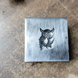 Könnte beinhalten: Ein Stempel aus silbernem Metall mit einem Eulenmotiv. Die Eule blickt nach vorne mit ausgebreiteten Flügeln. Der Stempel ist rechteckig und hat eine glatte Oberfläche.