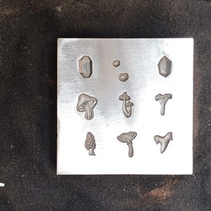 May include: A metal stamp set with nine different designs, including mushrooms, crystals, and other nature-inspired shapes. The stamps are arranged in a grid on a silver metal plate.