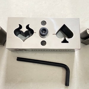 Puede incluir: Un bloque de metal con tres recortes: un corazón con llamas, una pica y un espacio en blanco. También se muestra una pica de metal y un corazón de metal con llamas. Se incluye una llave Allen negra.