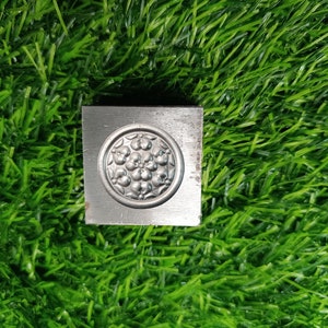 May include: A silver metal stamp with a circular design of a flower with multiple petals and a center filled with small circles. The stamp is on a green grassy surface.