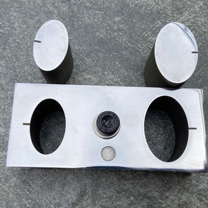 May include: A silver metal tool with two round holes and a small black knob in the centre. Two silver metal cylinders are placed on top of the tool.