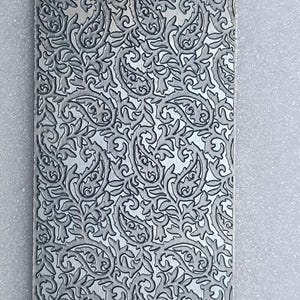 May include: A rectangular, silver-coloured metal plate with a detailed, embossed paisley pattern. The design features swirling leaves and floral elements, creating a textured surface. The plate is approximately 15 cm long and 7.5 cm wide.