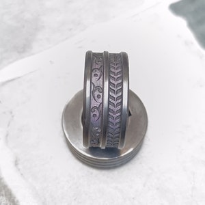 May include: A silver ring with two decorative bands. One band features a floral design, and the other has a leaf pattern. The ring is set on a silver, circular base, and the background is a light gray and white tile.