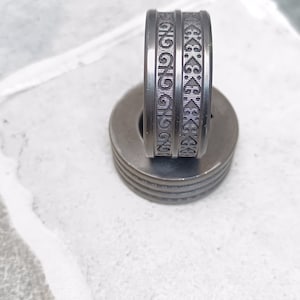 Könnte beinhalten: Ein silberfarbener Ring mit einem eingravierten Muster aus Herzen und Wirbeln. Der Ring liegt auf einem Stapel aus Metallscheiben. Das Design des Rings ist symmetrisch und detailliert.