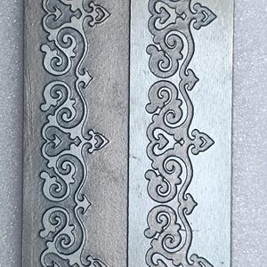 May include: A close-up of a silver metal sheet with a decorative pattern etched into the surface. The pattern is a repeating design of swirls and leaves.
