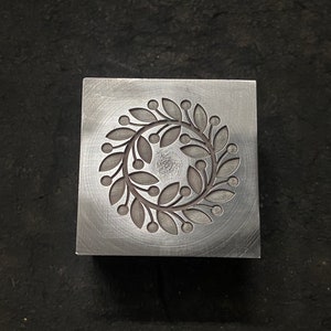 Puede incluir: Un bloque de metal plateado con un diseño circular de una corona de hojas y bayas.