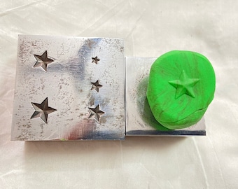 May include: Two metal blocks, one with star cutouts of varying sizes, the other with a green, star-shaped impression. The metal has a brushed, industrial finish. The green material appears to be clay or a similar moldable substance.