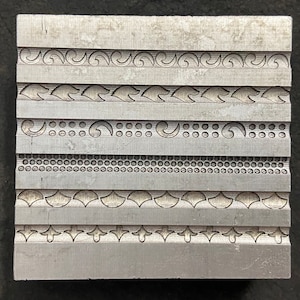 May include: A rectangular metal block with etched decorative patterns. The block features horizontal bands with various designs, including swirls, dots, and leaf-like shapes. The metal has a silver colour and a matte finish.
