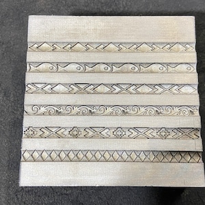 Op de afbeelding: Een metalen plaat met zes rijen verschillende geëmbosseerde ontwerpen. De ontwerpen omvatten geometrische patronen, vissen en bloemmotieven.