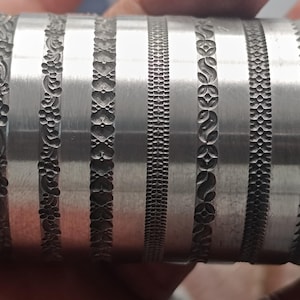 Könnte beinhalten: Eine Metallrolle mit mehreren dekorativen Mustern, die in ihre Oberfläche graviert sind. Die Rolle wird verwendet, um Prägemuster auf verschiedenen Materialien zu erstellen.