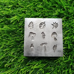 May include: A metal mold with various designs for crafting, including mushrooms, leaves, and a monstera leaf. The mold is gray and has a textured surface.