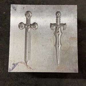 May include: A metal plate with two sword designs embossed into the surface. The swords are both silver in color and have intricate details.