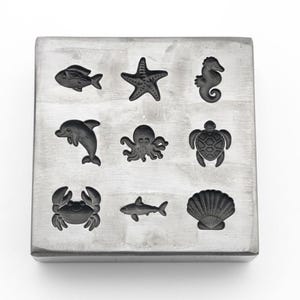 May include: A square, silver-coloured metal mould featuring nine sea creature designs. The designs include a fish, starfish, seahorse, dolphin, octopus, turtle, crab, shark, and seashell. The mould is on a white background.