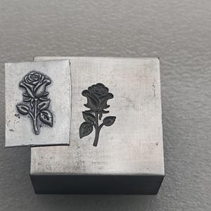 May include: A square metal stamping tool featuring a recessed rose design, positioned next to a small rectangular metal plate that displays a raised, embossed rose impression created by the tool. Both items are made of a smooth, silver-toned metal.