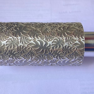 May include: A silver rolling pin with a leaf pattern embossed on the surface. The rolling pin has a handle on each end.