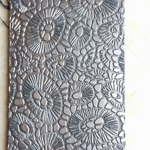 May include: A silver-colored embossed sheet of paper with a repeating pattern of small circles and larger circles with radiating lines.