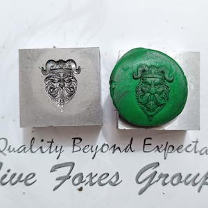 May include: A close-up of a metal stamp with a design of a face with horns and a crown. The stamp is used to create an impression in green clay.