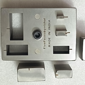 Op de afbeelding: Een set zilverkleurige metalen componenten, waaronder een rechthoekige basis met uitgesneden secties en verschillende kleinere, rechthoekige stukken. De tekst "Five Foxesbyvishal MADE IN INDIA" staat op de basis. De componenten lijken precisie bewerkt.