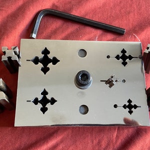 May include: A metal plate with four decorative cutouts and a center hole. There are four metal blocks with matching cutouts, used for creating decorative metalwork.