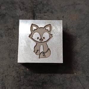 May include: A metal stamp with a cartoon fox design. The fox is brown with a white face and a white tail.