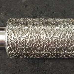 以下が含まれることがあります： 表面に花柄が刻まれた銀色の金属ローラー。ローラーは両端に滑らかな円筒形のハンドルが付いています。