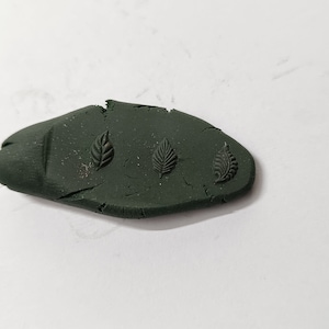 May include: A dark green, oval-shaped piece of clay with three leaf designs pressed into the surface.