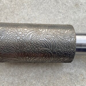 Könnte beinhalten: Ein silberner Nudelholz mit einer strukturierten Oberfläche, die ein Muster aus Blättern und Kreisen zeigt. Das Nudelholz ist aus Metall und hat einen glatten Griff.