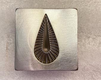May include: A metal stamp with a teardrop-shaped design. The teardrop has a textured, radiating pattern.