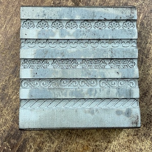 Könnte beinhalten: Ein Metallblock mit fünf Reihen verschiedener dekorativer Muster, die in die Oberfläche graviert sind. Der Block ist grau und hat eine leicht strukturierte Oberfläche.