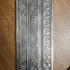 May include: A silver metal plate with three different embossed patterns. The patterns are a series of triangles, a Greek key design, and a series of curved lines.