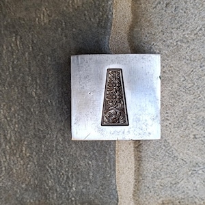 Puede incluir: Un cuadrado de metal plateado con un diseño decorativo en el centro. El diseño es un rectángulo vertical con detalles intrincados.