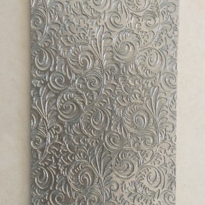 Könnte beinhalten: Eine silberne Metallplatte mit einem geprägten Blumenmuster. Das Muster besteht aus Spiralen und Blättern.