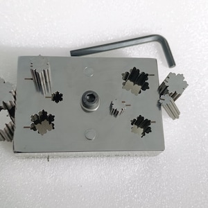 May include: A metal tool with a rectangular base and multiple star-shaped punch dies. The dies are arranged in a grid pattern and can be removed for use. The tool also includes a small wrench for tightening the dies.