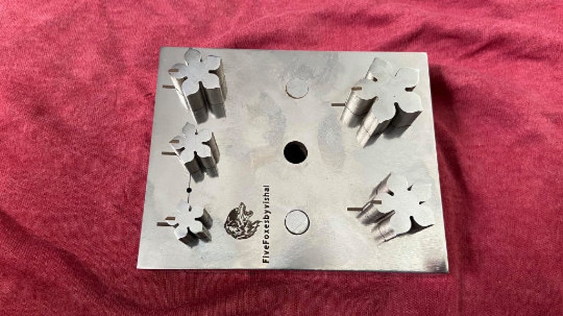 May include: A metal punch set with five different flower-shaped punches. The punches are arranged in a grid pattern on a metal plate. The plate is labeled "FiveFoxesbyVishal".