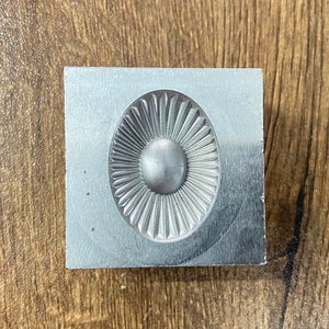 May include: A silver metal mold with a raised, circular design. The design features a central circle surrounded by radiating lines.