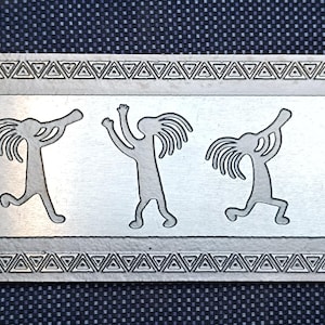 May include: A silver metal bookmark with a decorative design of five stylized figures with long hair and arms raised. The figures are arranged in a row and are surrounded by a border of triangles.