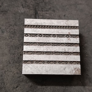 May include: A silver metal block with six rows of decorative patterns embossed on the surface.