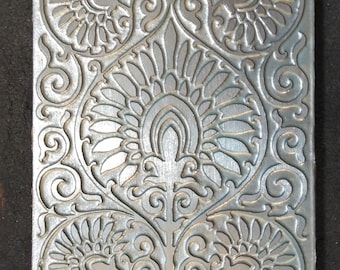 May include: A silver metal tile with an embossed design of repeating floral patterns. The tile has a textured surface and a slightly raised design.
