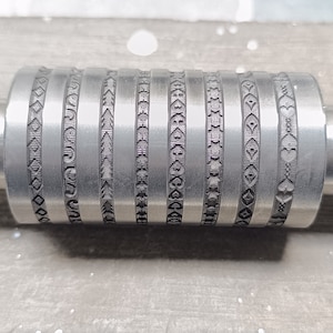 Könnte beinhalten: Ein zylindrisches Metallwerkzeug mit aufwendigen geätzten Mustern. Das Werkzeug hat einen zentralen Zylinder mit vertikalen Bändern aus Mustern, darunter Herzen, Sterne und geometrische Formen. Die Enden sind massiv und zylindrisch, wobei ein Ende eine Kerbe aufweist.
