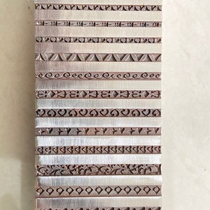 Könnte beinhalten: Ein Metallblock mit 20 verschiedenen gravierten Mustern, darunter Punkte, Linien, Herzen und Spiralen. Der Block ist silberfarben mit einer gebürsteten Oberfläche.